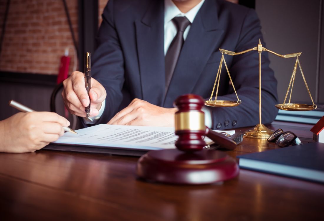 A lawyer and client signing an agreement on a desk, next to a gold weighing scale of justice and a judge gavel as they discuss legal debt collection for the client.  